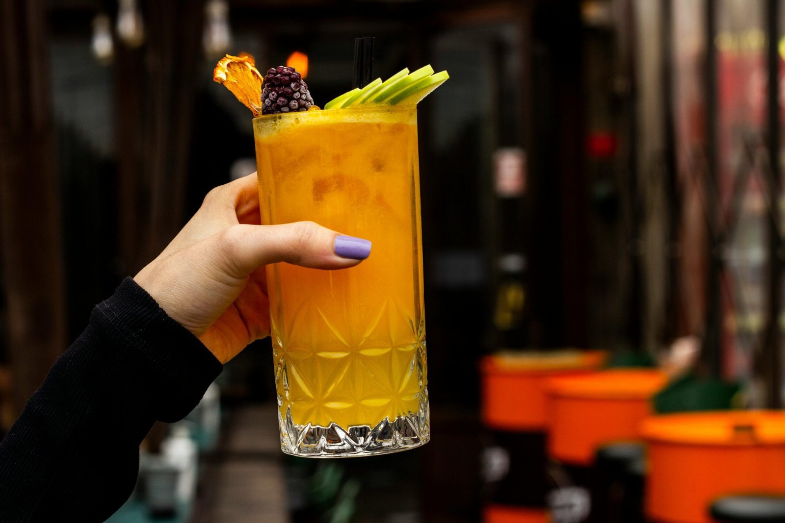A stylish orange cocktail garnished with fruit, held by a woman with purple nails, ideal for lifestyle and beverage themes.